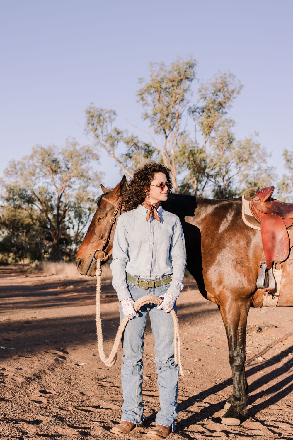 Person in denim and light shirt stands next to saddled brown horse in outback setting.
