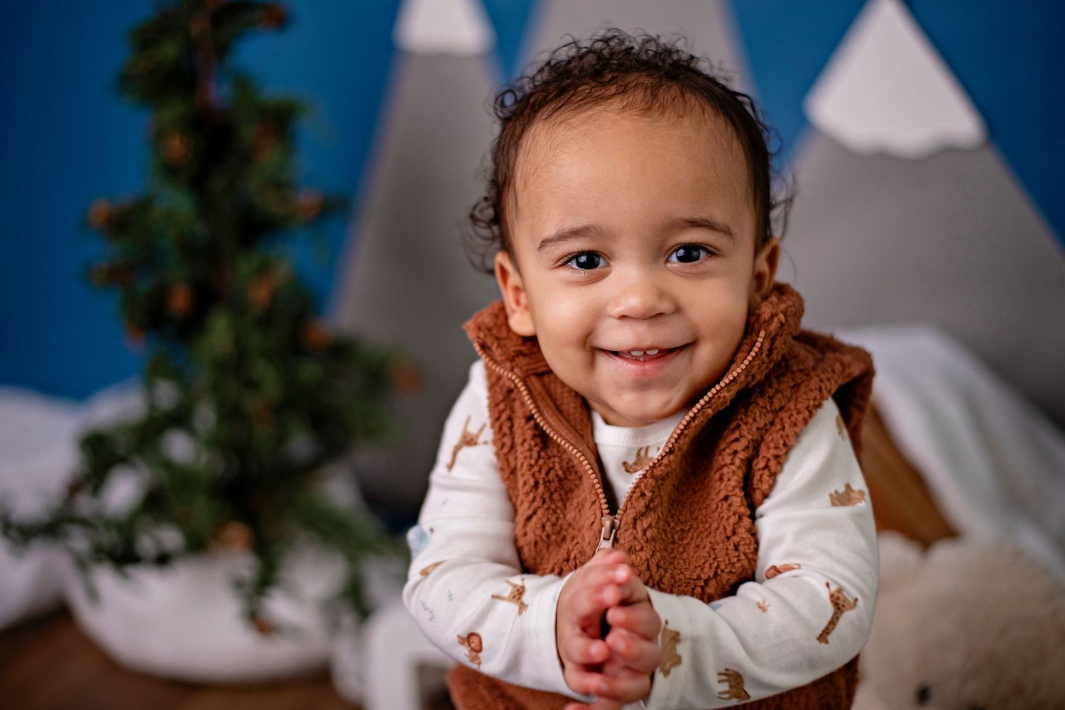 Baby wearing brown fleece vest and white shirt sits in front of teepee and holiday decor in studio setting.