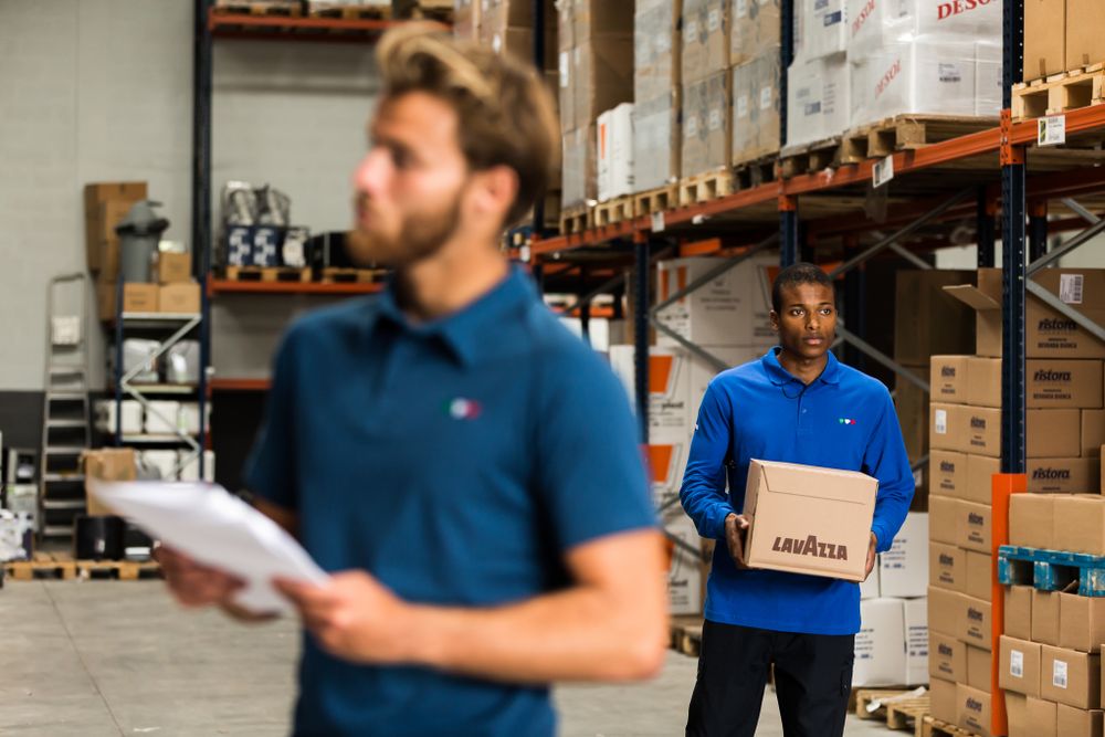 Two warehouse workers in blue polo uniforms reviewing inventory among storage shelves stacked with boxes.