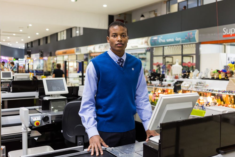 Retail worker in blue vest and white shirt standing at checkout counter in supermarket.
