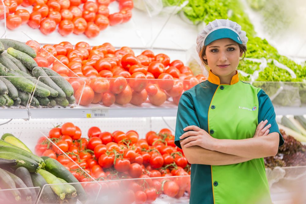 Produce worker in green uniform standing by fresh vegetables and tomatoes in grocery store display.