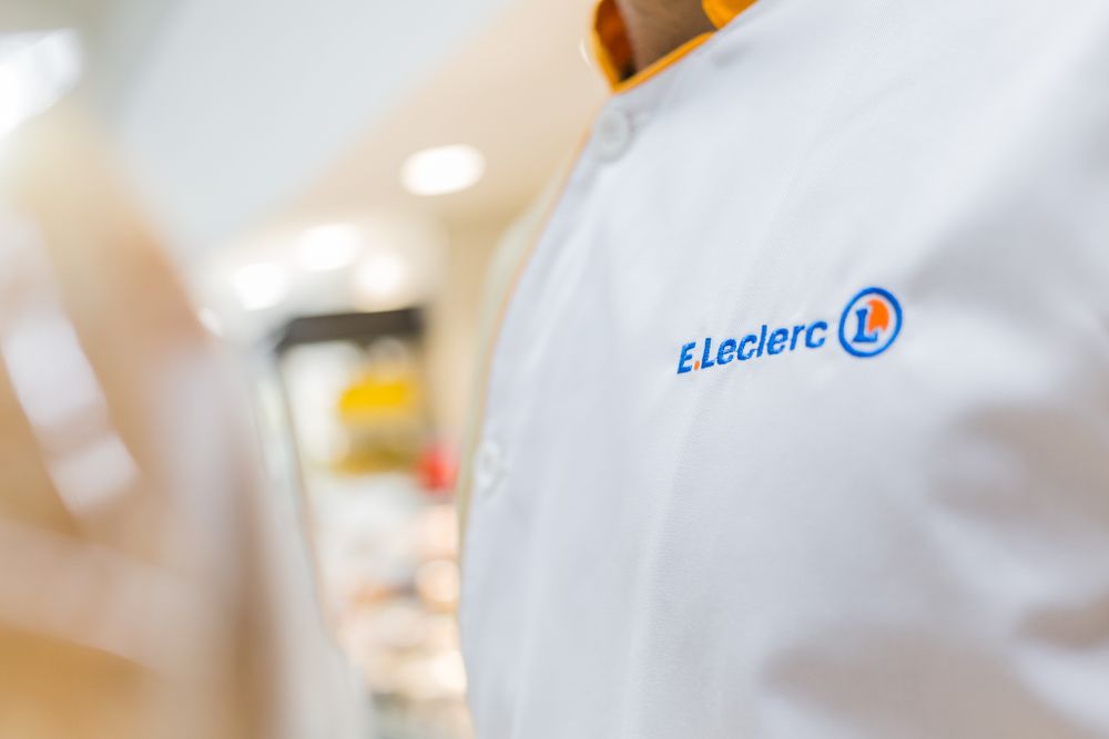 Close-up view of a white E.Leclerc uniform shirt with blue embroidered logo in a retail store setting.