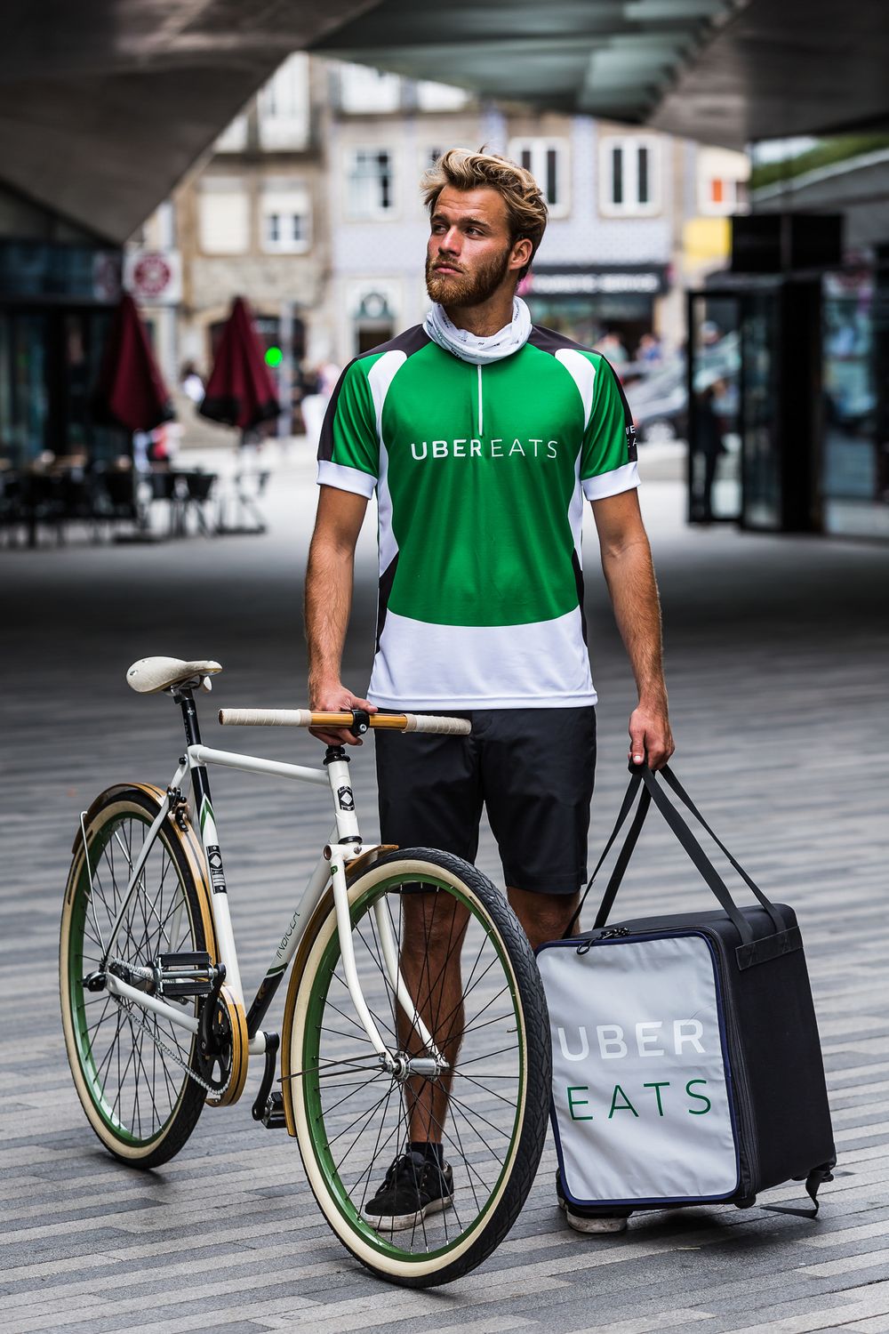 An Uber Eats delivery person in green uniform stands with a bicycle and insulated delivery bag on a city street.