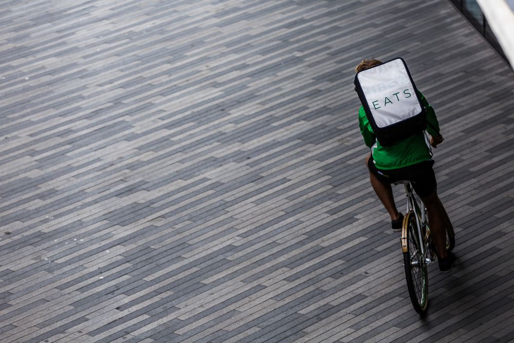 A food delivery cyclist with a green cubic backpack rides on a gray textured road surface.