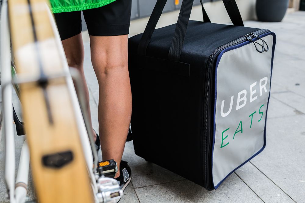 Black Uber Eats delivery bag sits on sidewalk next to driver's legs and bicycle in urban setting.