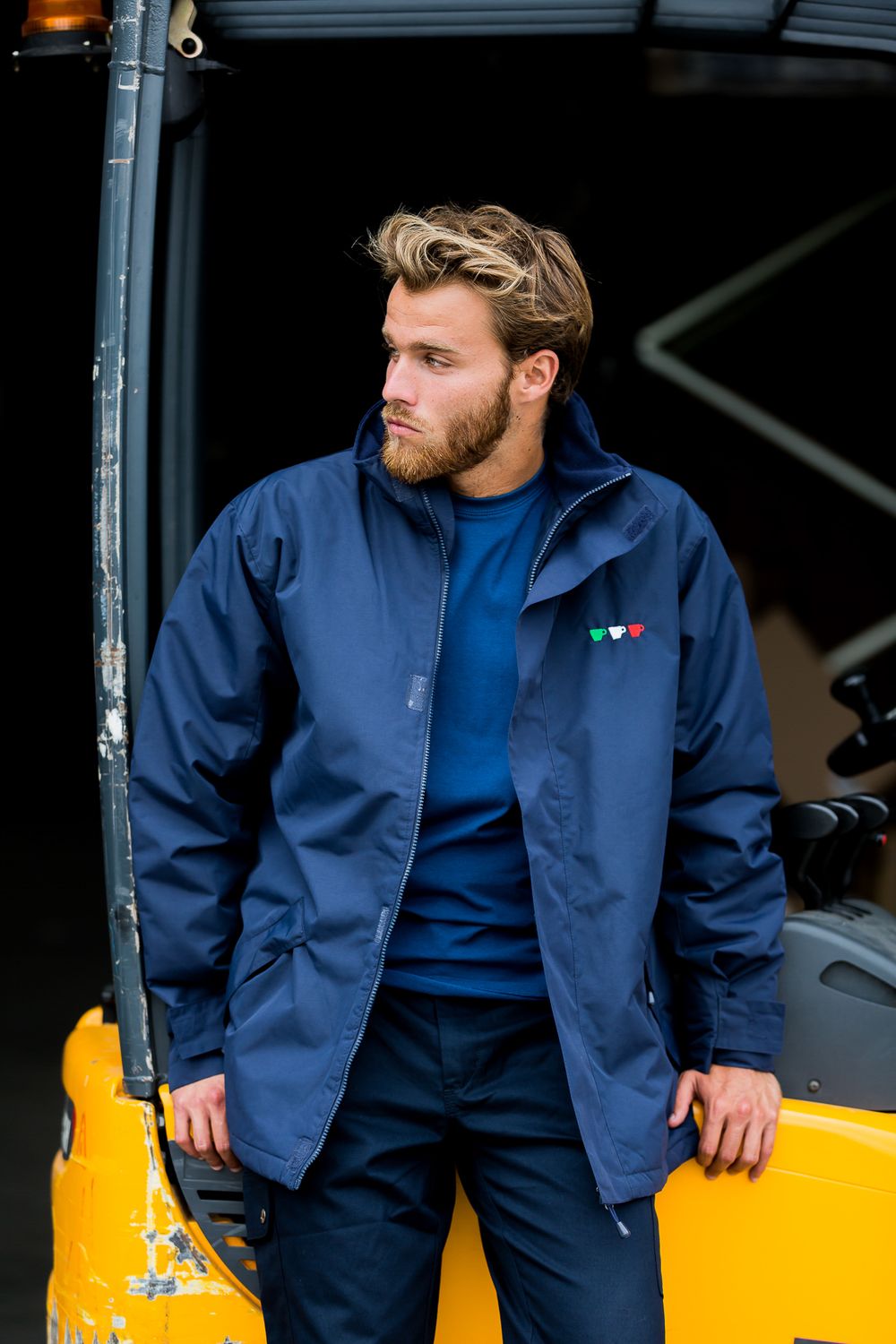 A person wearing a navy blue jacket and sweater poses against a yellow industrial equipment background.