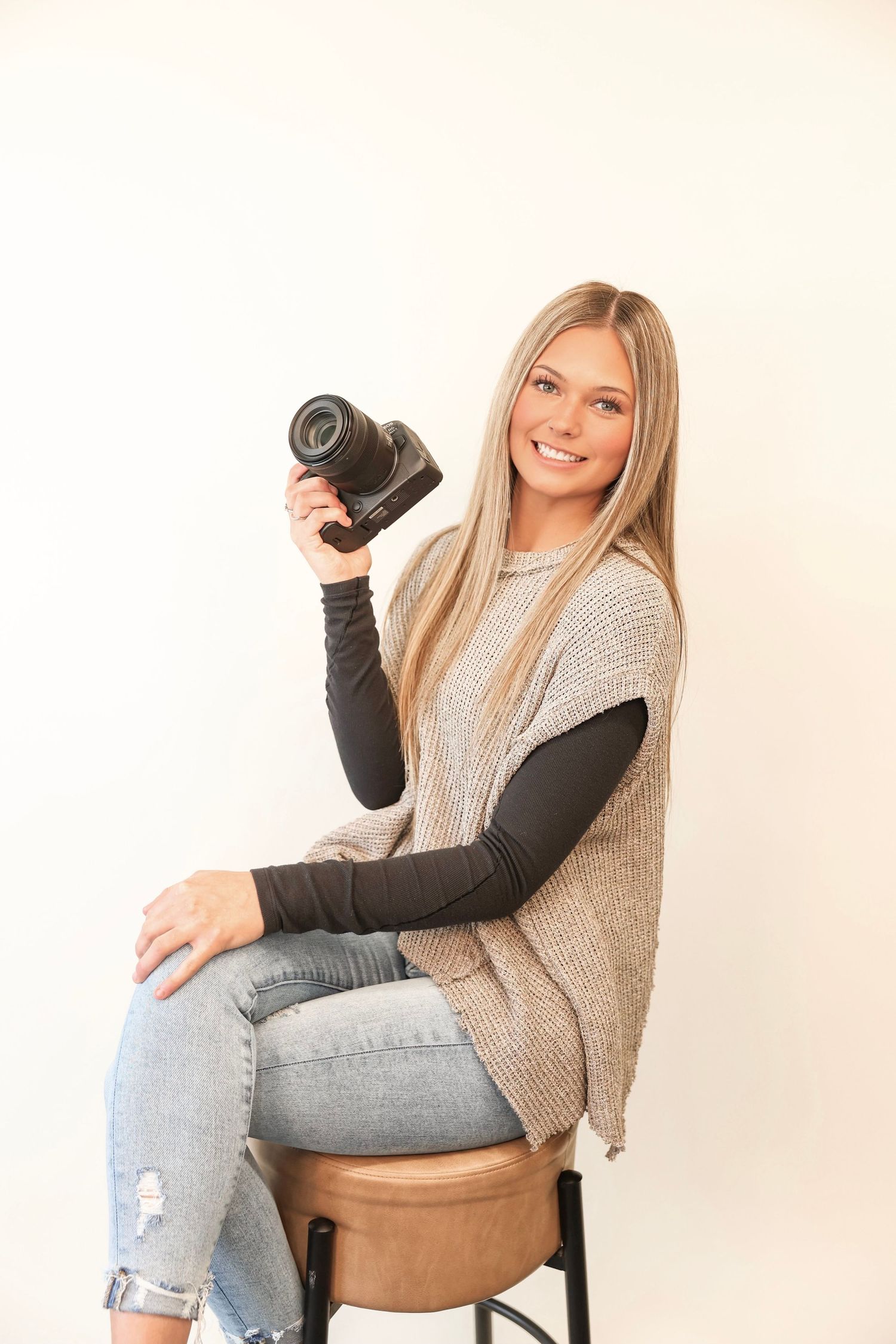 Photographer wearing tan sweater vest and black long sleeve holds camera while sitting on brown leather stool.