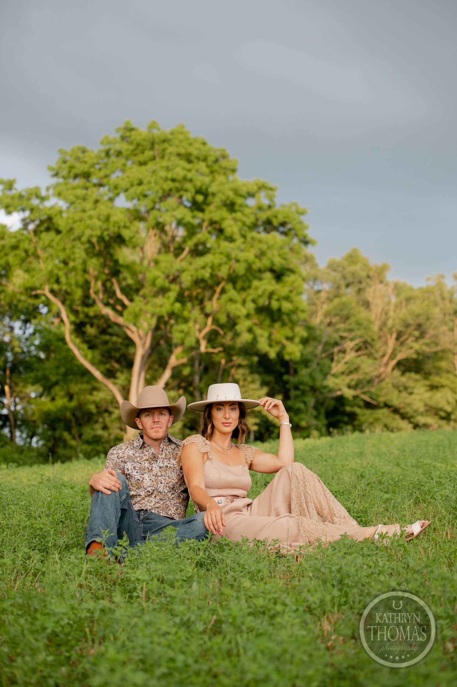 A western-styled couple sitting on a grassy hillside during golden hour.