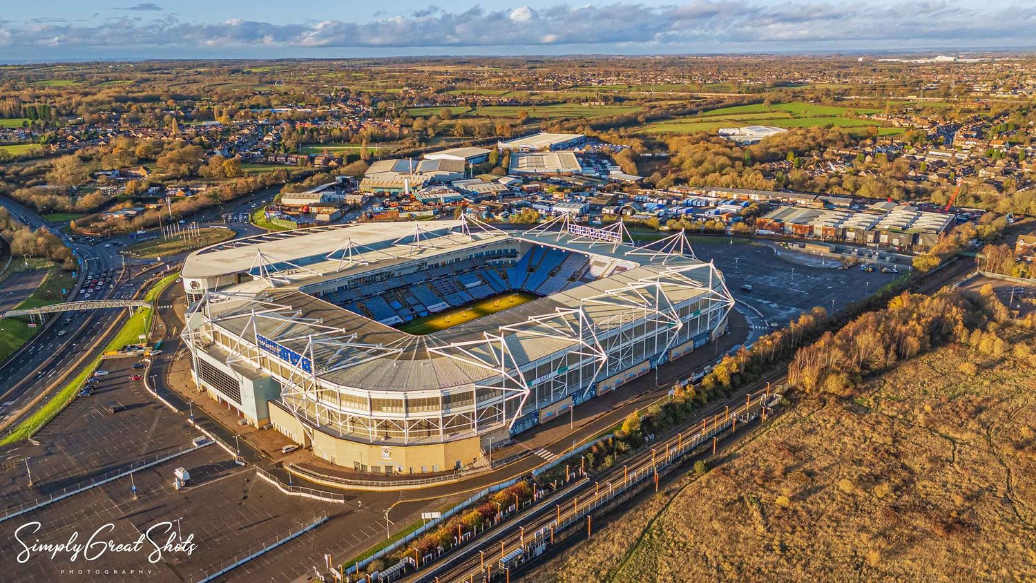 Aerial view of the Coventry Building Society Arena stadium surrounded by autumn trees and railway tracks under blue skies.