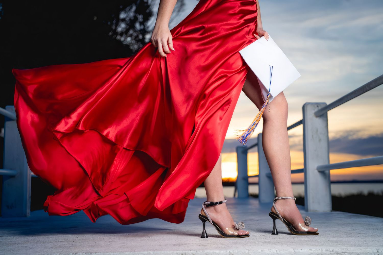 A flowing red satin dress billows in the breeze as high heels stand on a deck against a sunset sky.