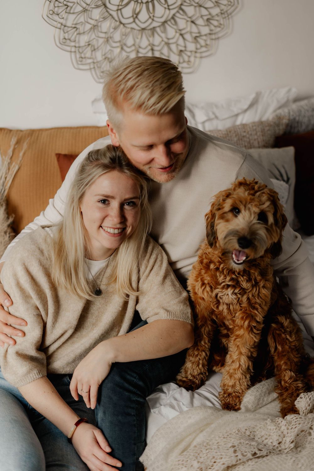 A cozy home scene with a happy couple in beige sweaters cuddling on a couch with their smiling goldendoodle dog.