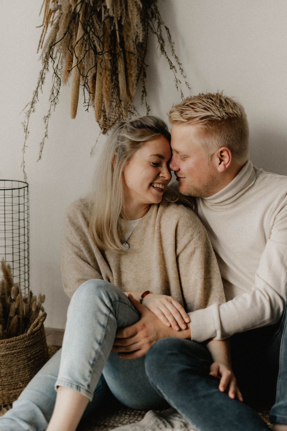 A couple in cozy cream sweaters share an intimate moment while sitting together against a neutral wall with dried flowers.