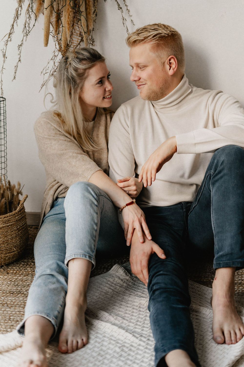 Couple in cozy cream sweaters sitting close together on floor with dried pampas grass decor in background.