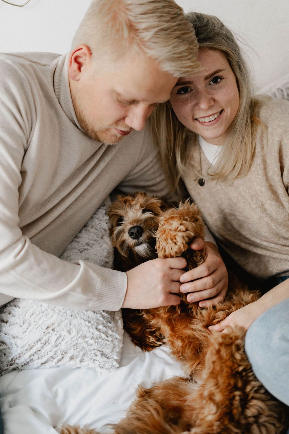 A couple in matching beige sweaters cuddles with their small fluffy dog on a cozy bed.