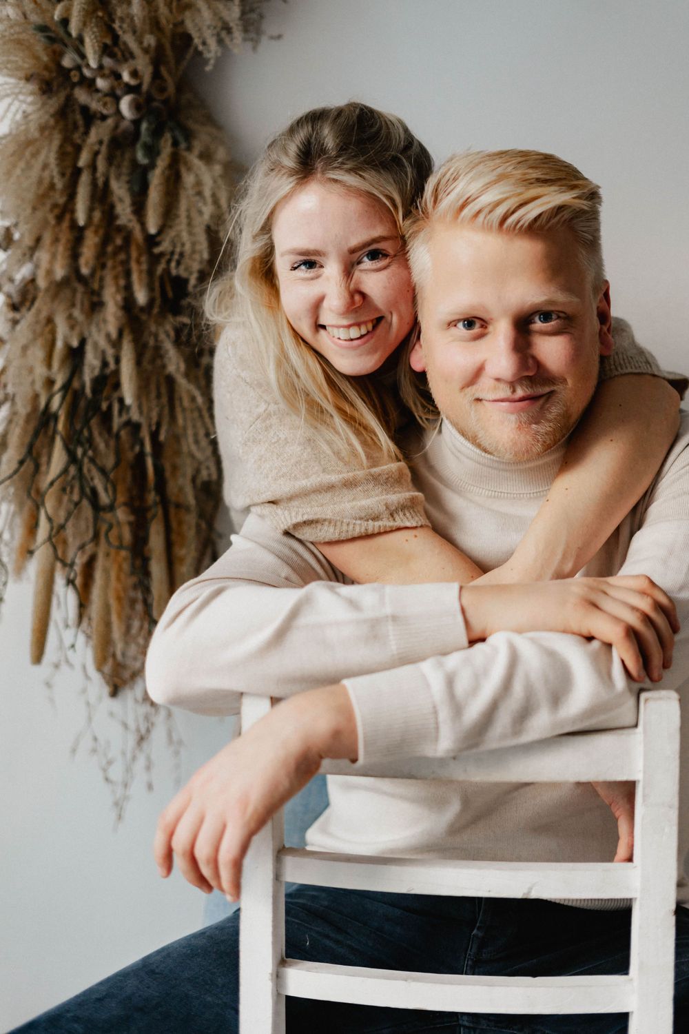 A couple in cozy cream sweaters embrace while sitting on a chair with dried pampas grass decor in the background.