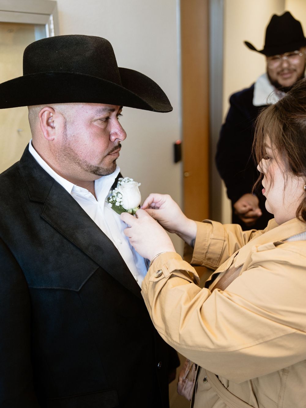 Someone in a black cowboy hat and formal western attire gets a white boutonniere pinned to their lapel before an event.
