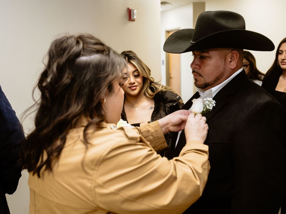 A person adjusts a black bowtie on a man wearing a black cowboy hat in a hallway.