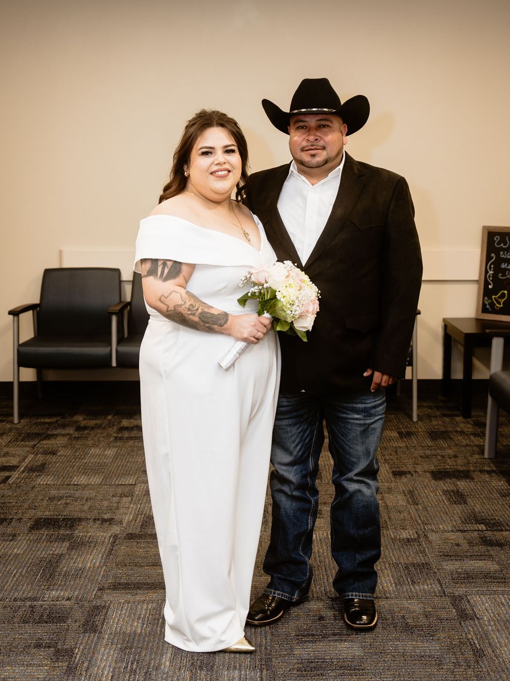 A bride in a white off-shoulder dress holding a bouquet stands with a groom wearing a black cowboy hat and jeans.