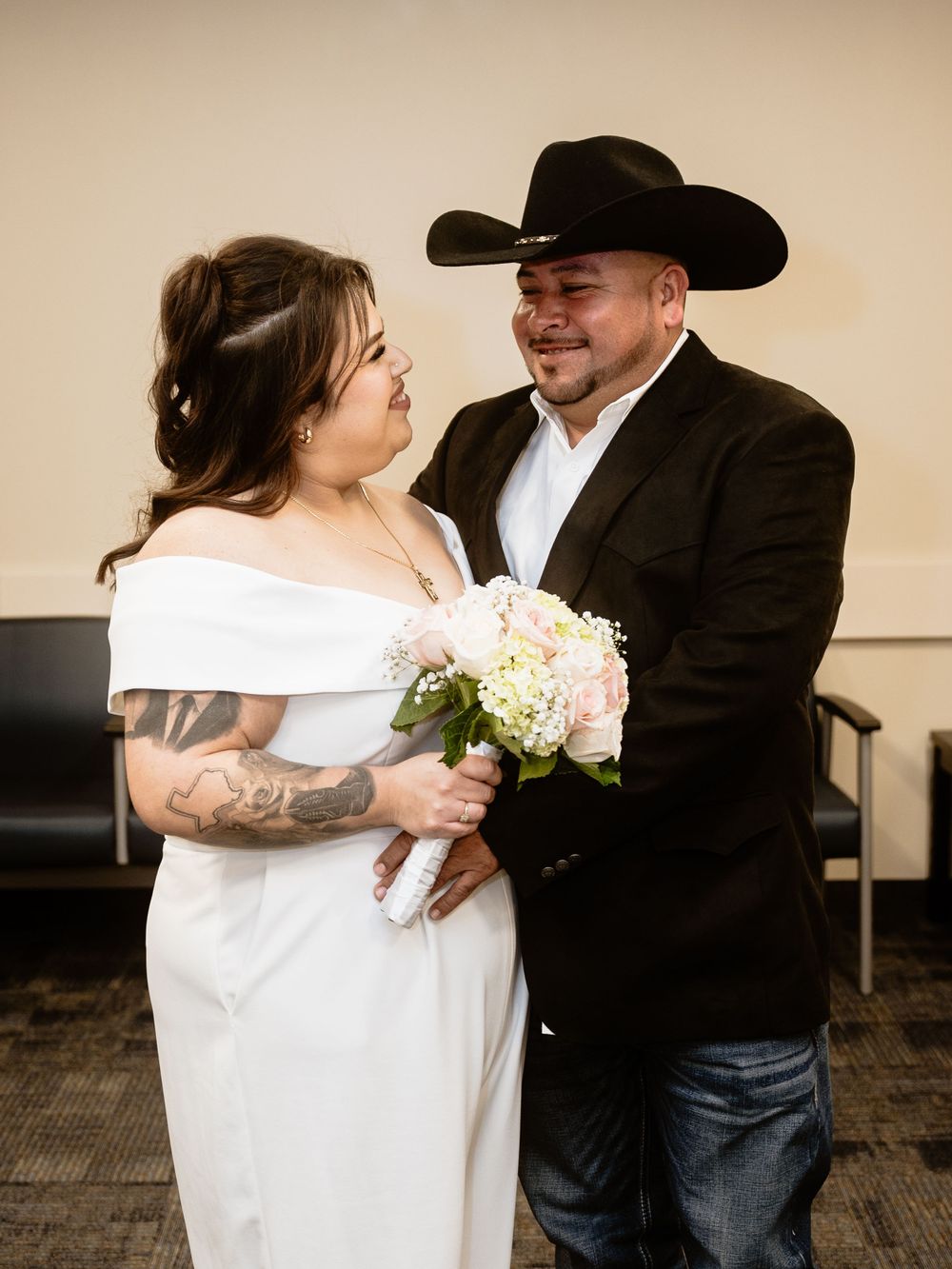 A bride in a white off-shoulder dress holds a blush bouquet next to her groom wearing a black cowboy hat and suit jacket.
