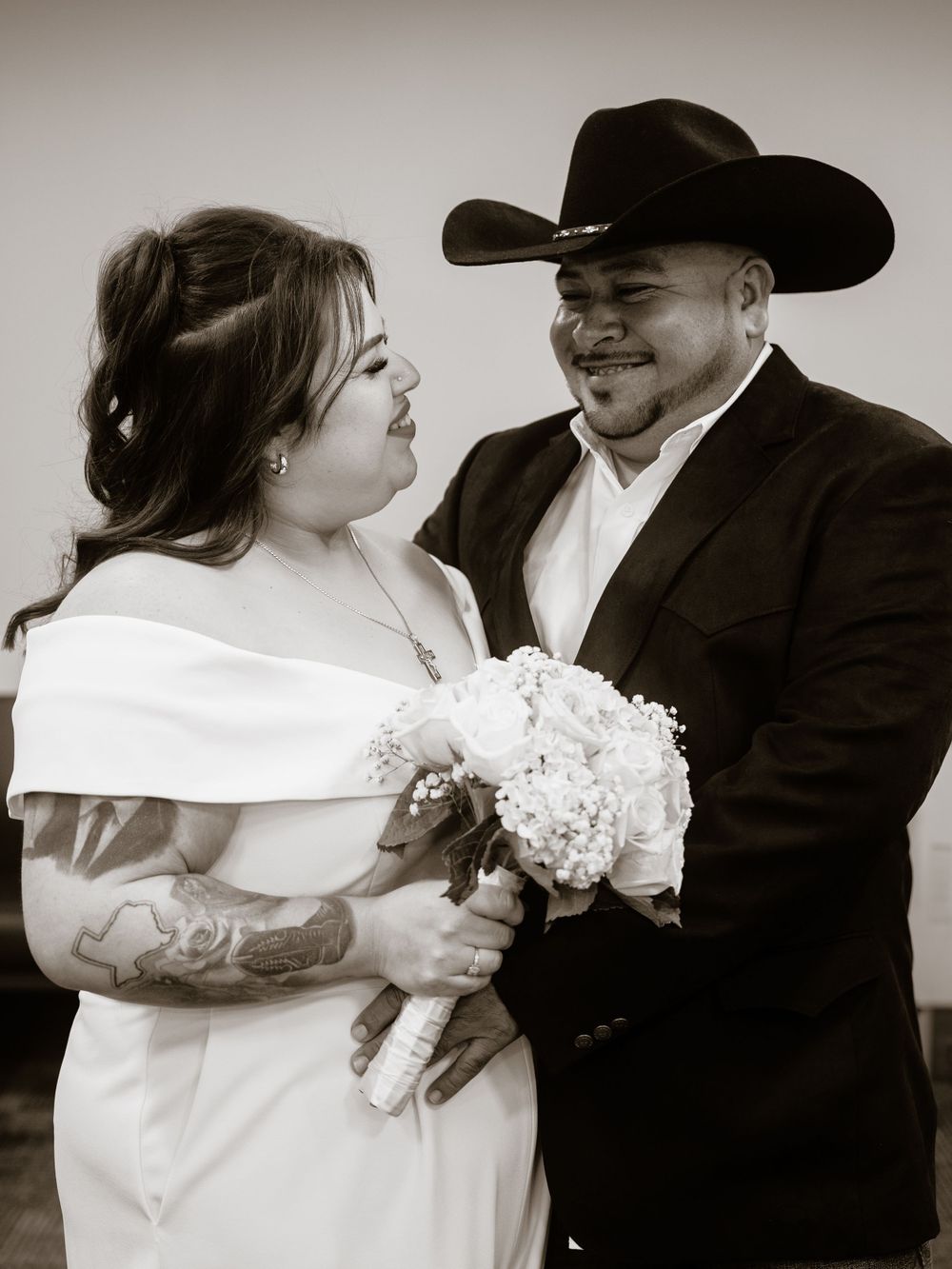 A black and white wedding portrait of a joyful couple, with the bride in white holding a bouquet and groom wearing a cowboy hat.