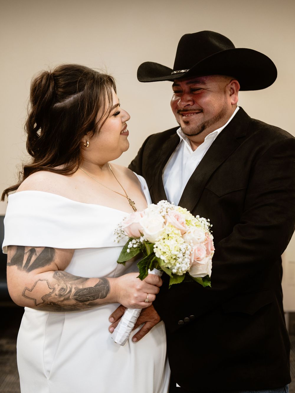 Newlywed couple share a joyful moment as bride in white dress holds bouquet and groom wears black cowboy hat.