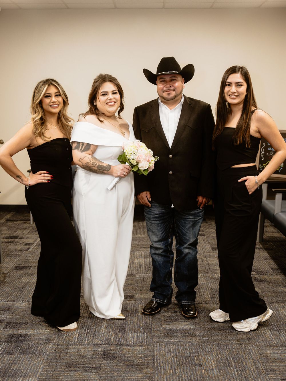 A group of four people pose together at a formal event, with women in black dresses and white gown, and a man in cowboy attire.