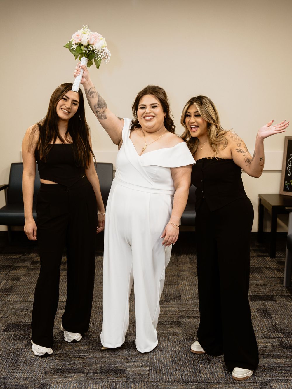 Three people celebrating at an event, with one person in white holding up a bouquet while flanked by two in black dresses.