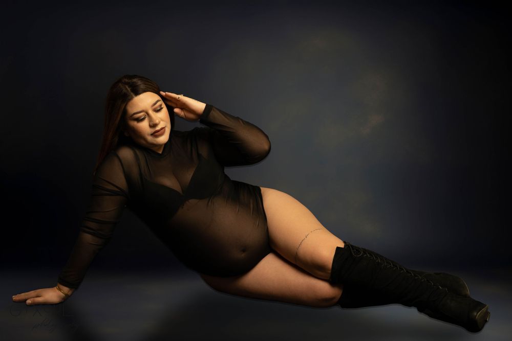 Dramatic maternity photo in black bodysuit and boots with moody lighting.