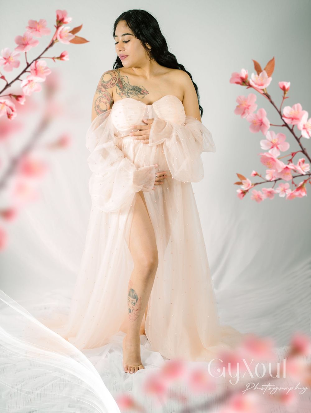 A maternity portrait with soft pink cherry blossoms features a flowing sheer dress and artistic composition.