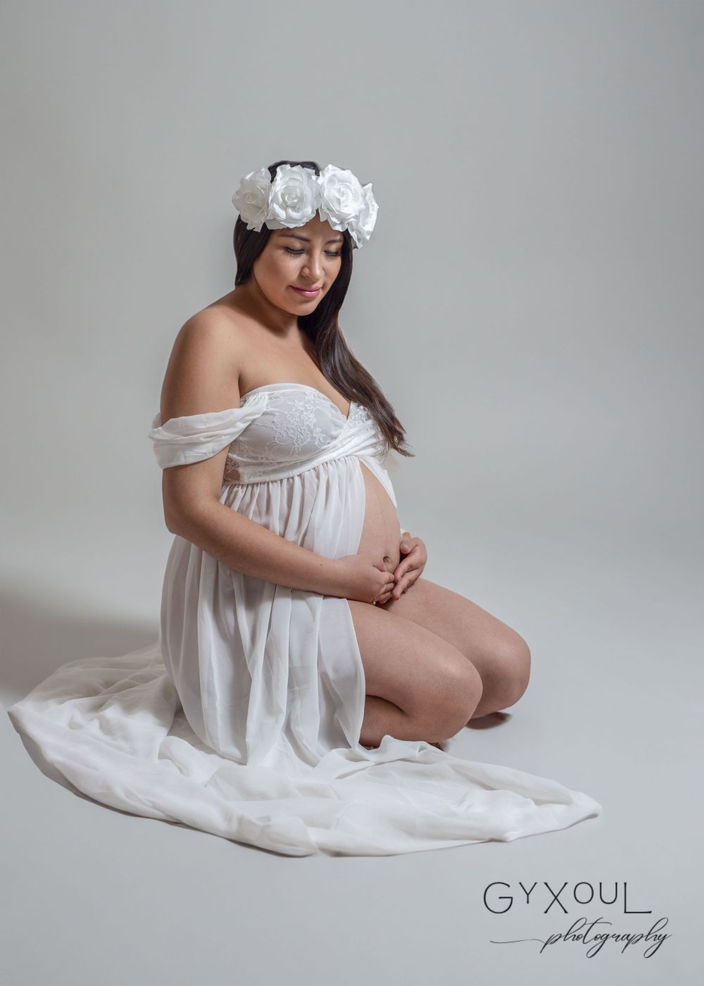 Ethereal maternity portrait with white lace dress and floral crown against grey backdrop.