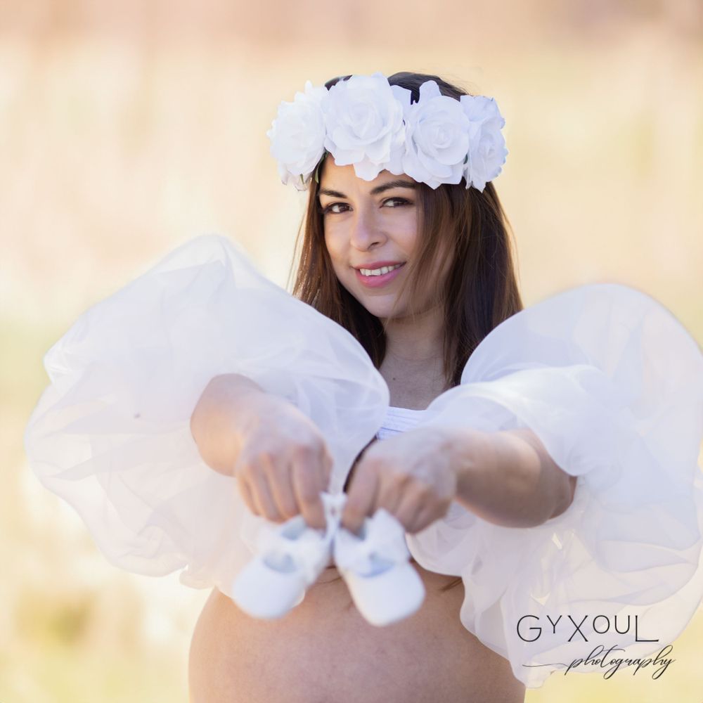 Sweet maternity moment captured holding tiny white baby shoes with floral crown.