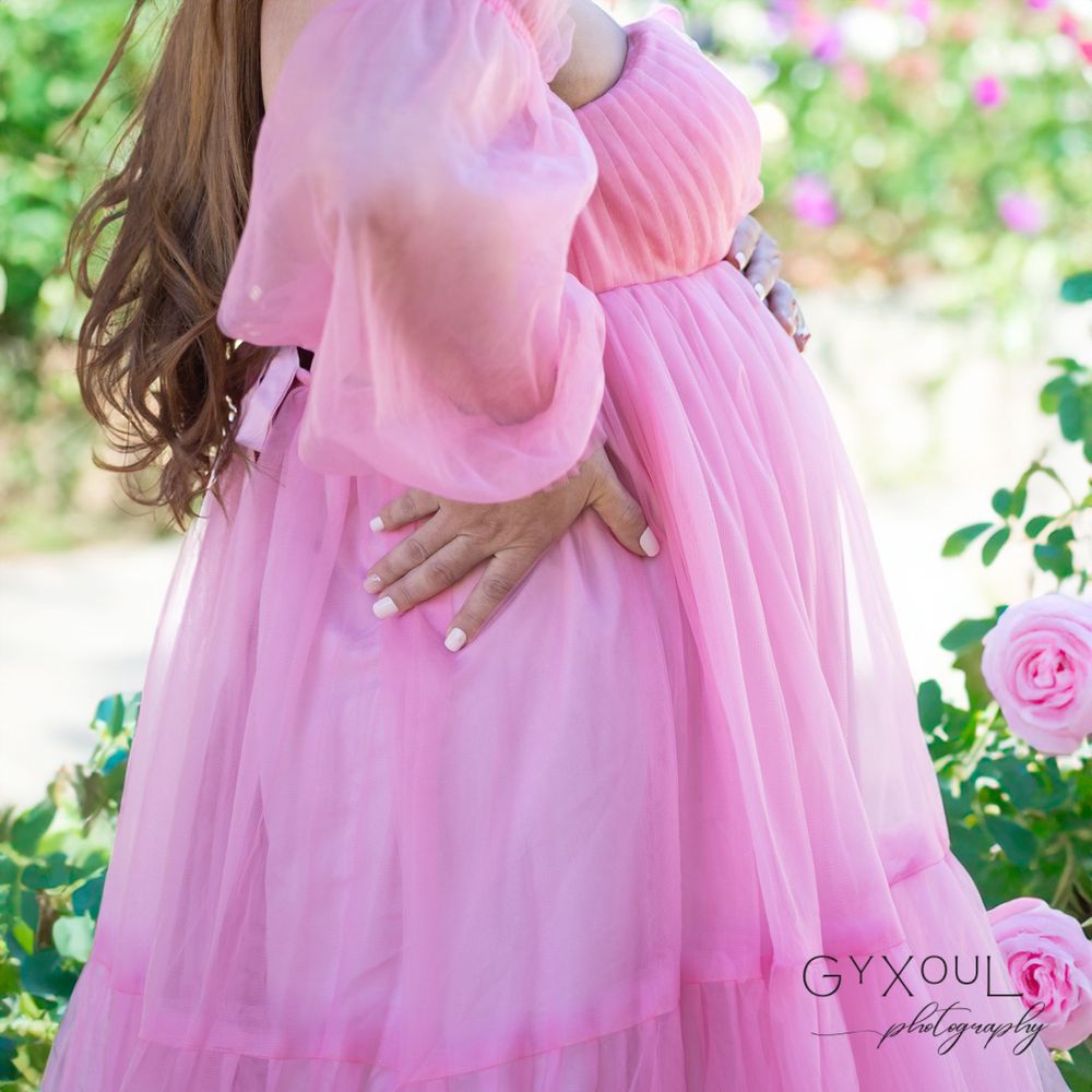Romantic outdoor maternity portrait in flowing pink gown surrounded by rose bushes.