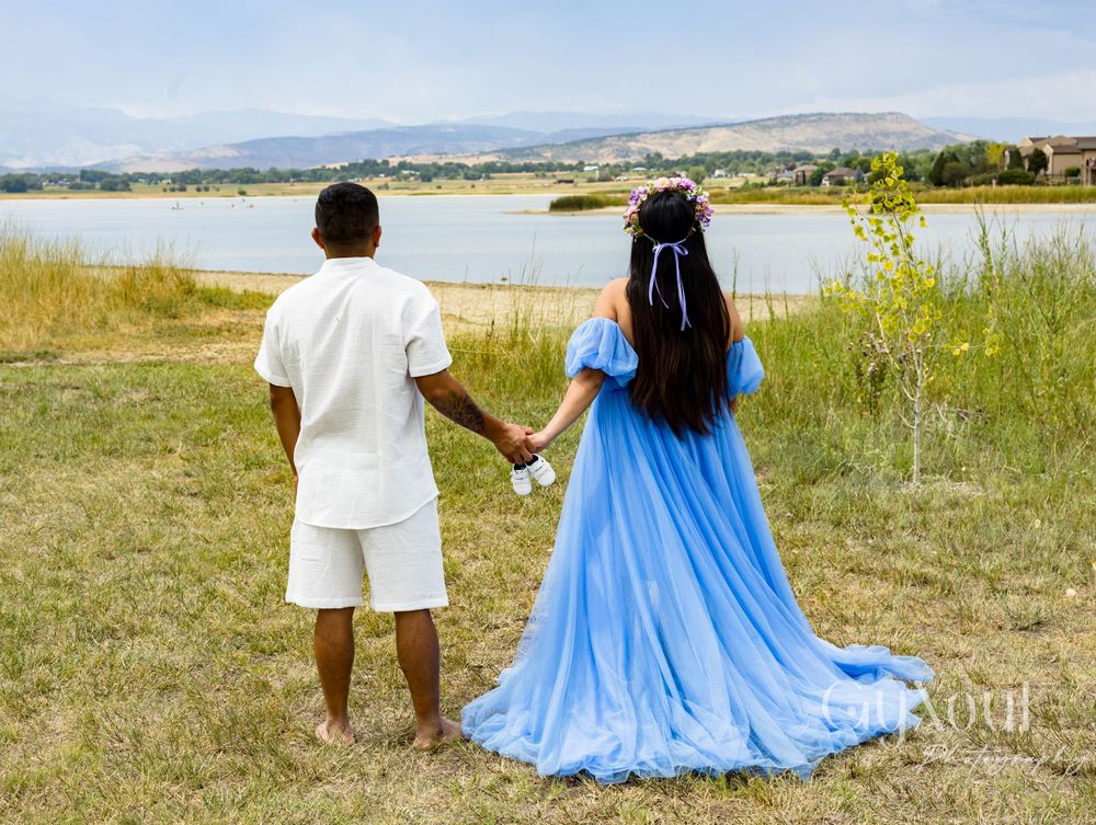A couple in white and blue stand holding hands overlooking a scenic lake and mountain landscape.