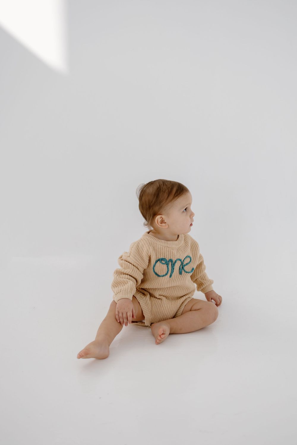 Baby in beige 'one' onesie sits on white background looking to the side.