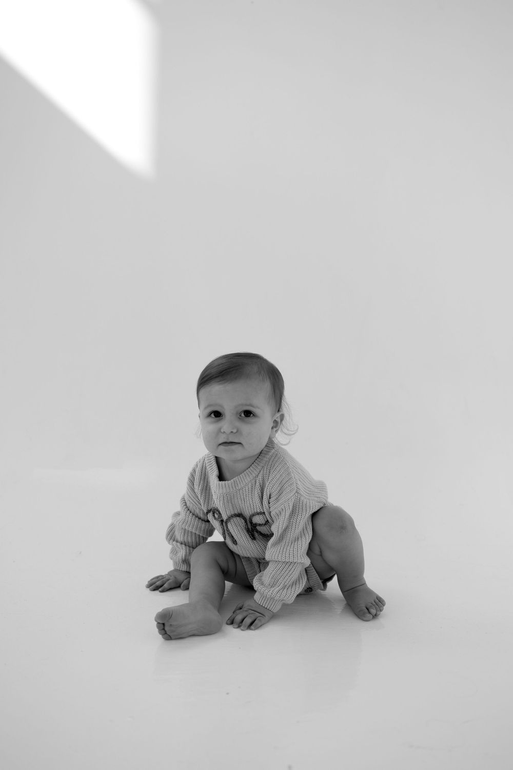Black and white photo of baby sitting on white backdrop.
