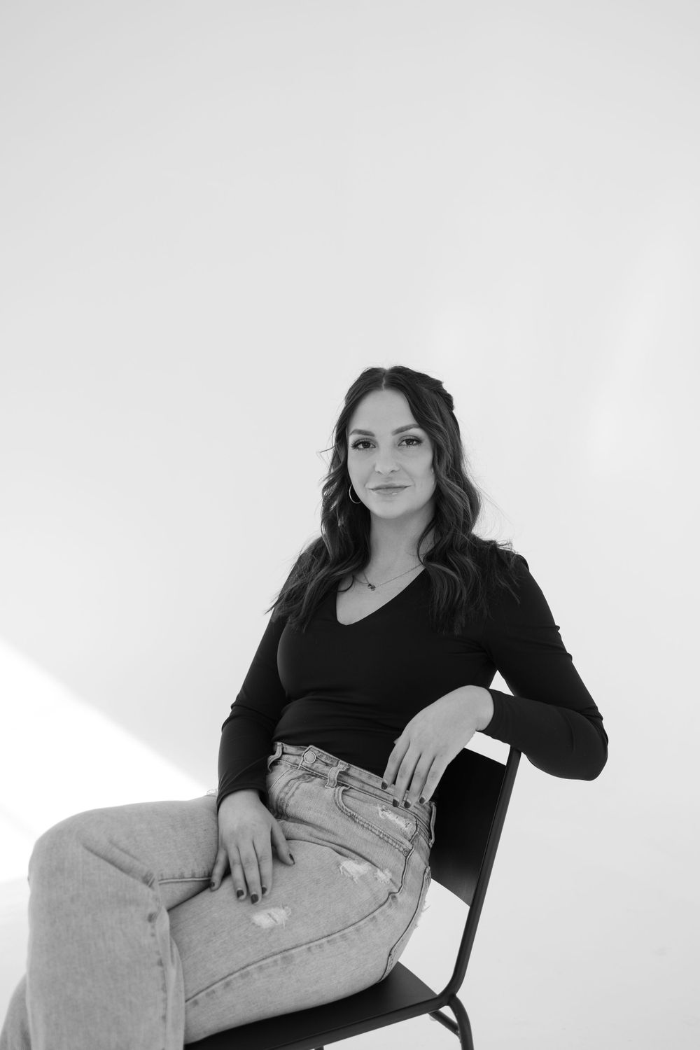 Woman in black top and light jeans poses on chair against white background.