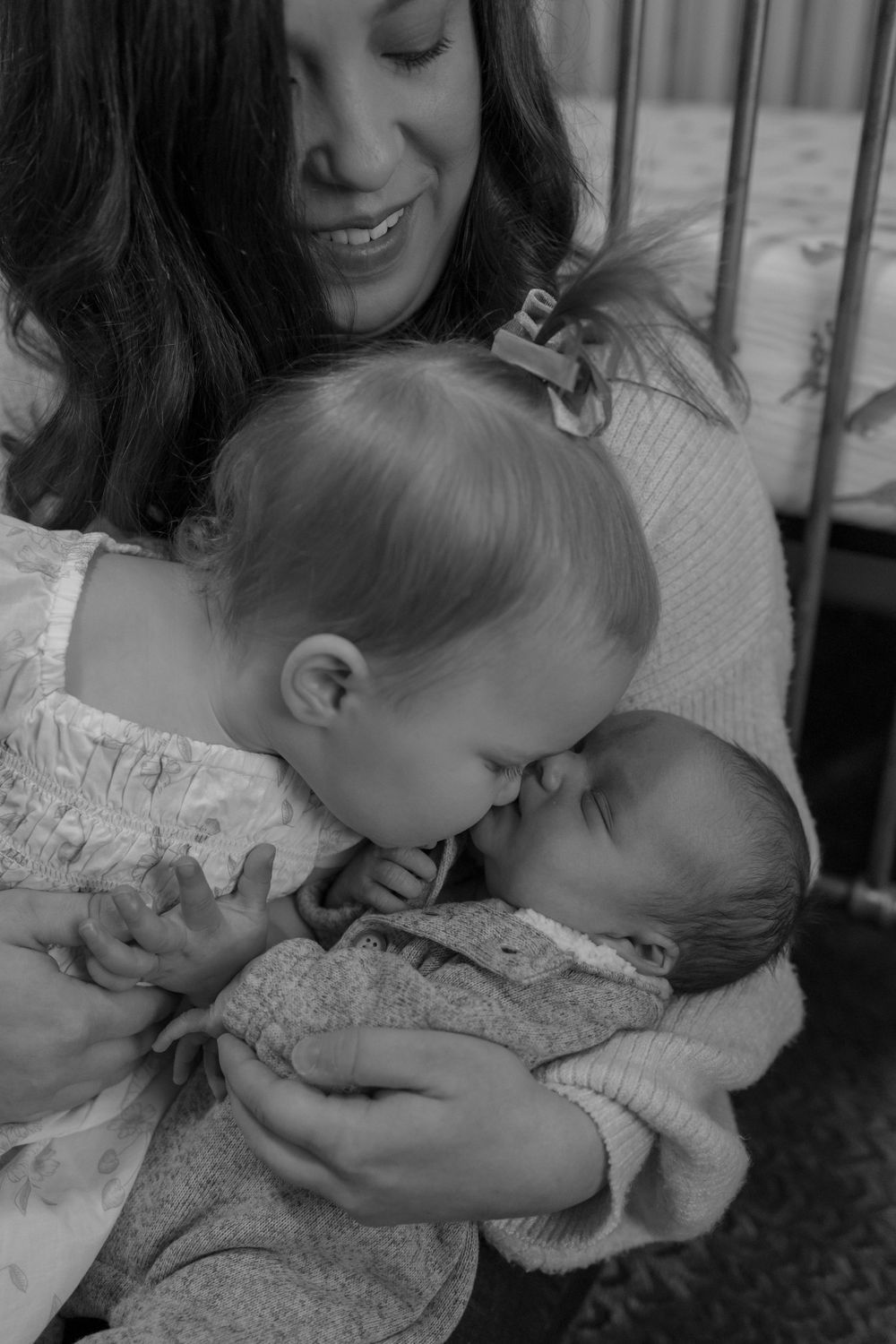 Tender black and white moment as toddler kisses newborn sibling while being held.