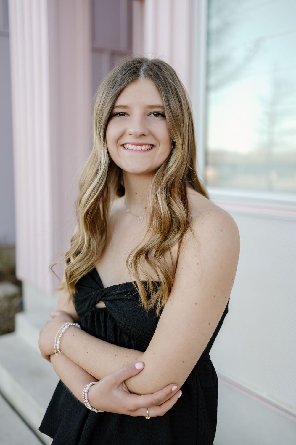 Young woman in black dress smiles warmly while posing against light colored building exterior.