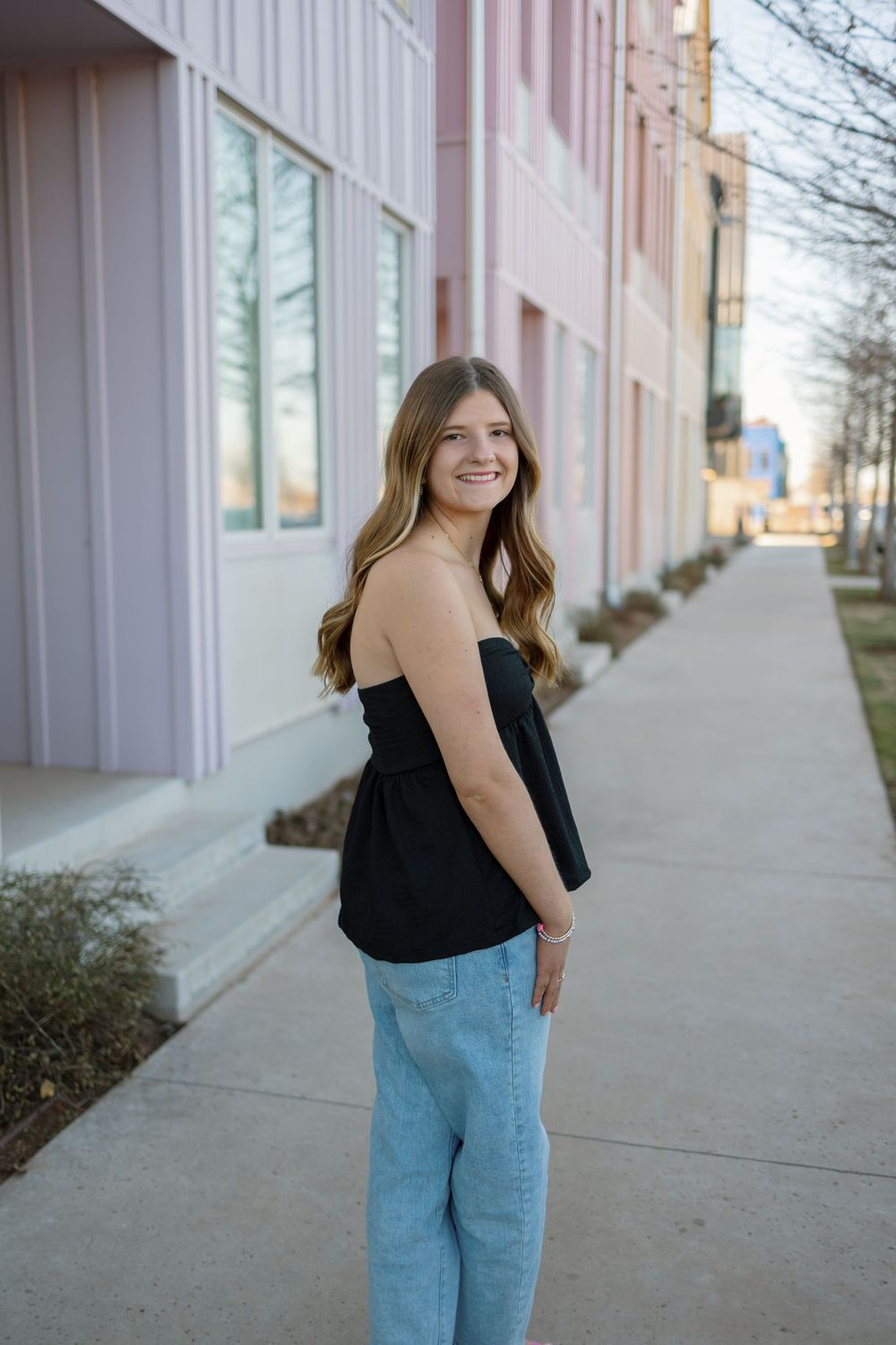 Woman in black top and jeans poses casually on sidewalk next to pastel colored building.