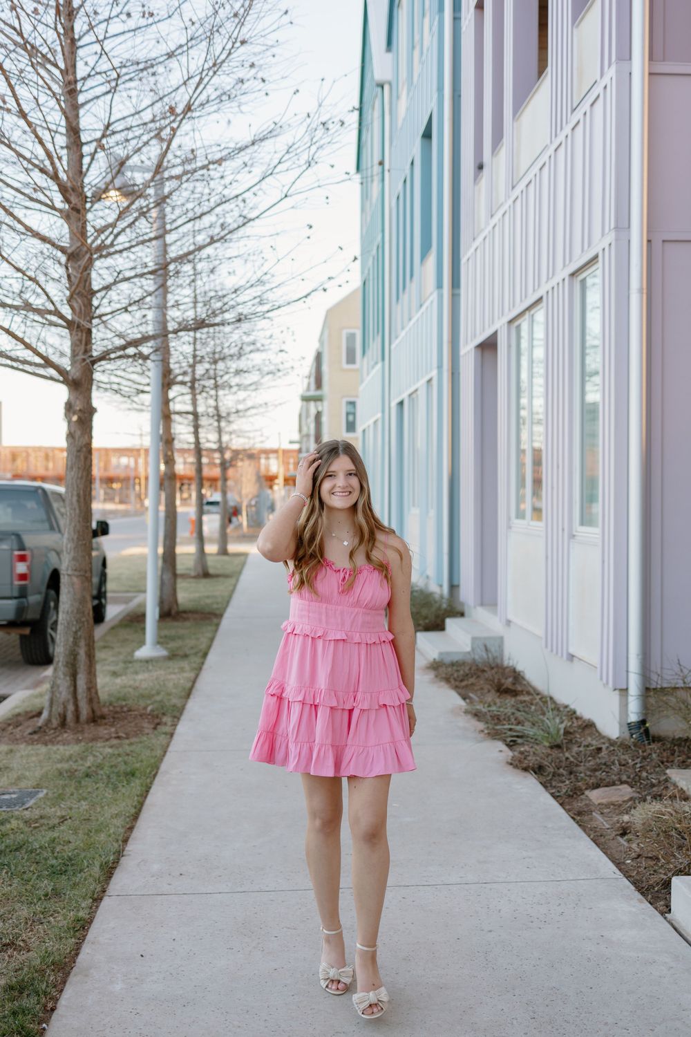 Woman in pink tiered dress poses on sidewalk near modern colorful building exterior.