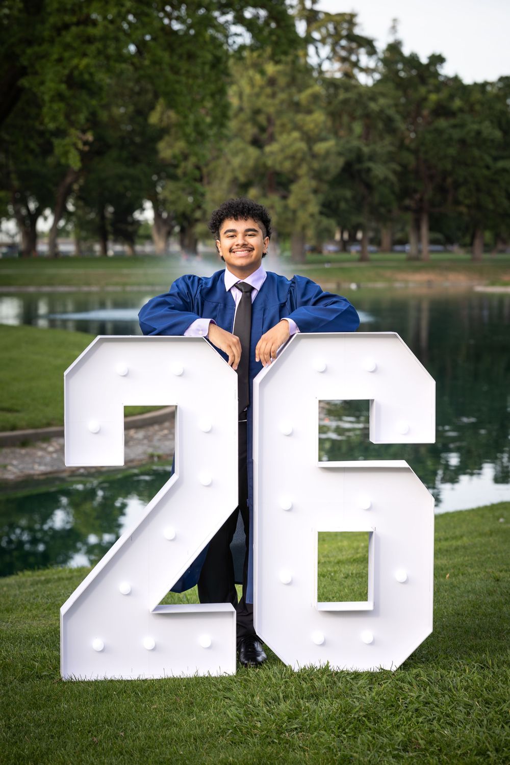 Young man in blue graduation gown smiling behind illuminated number 26 sign outdoors near pond.