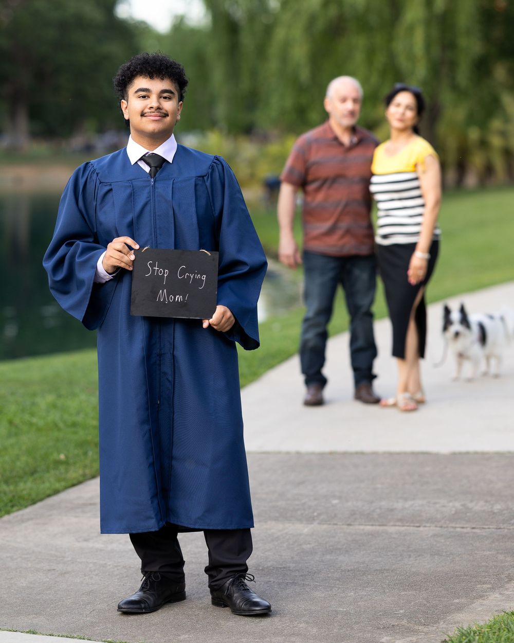 Graduate in blue cap and gown holds sign saying Stop Crying Mom with proud parents and dog in background.