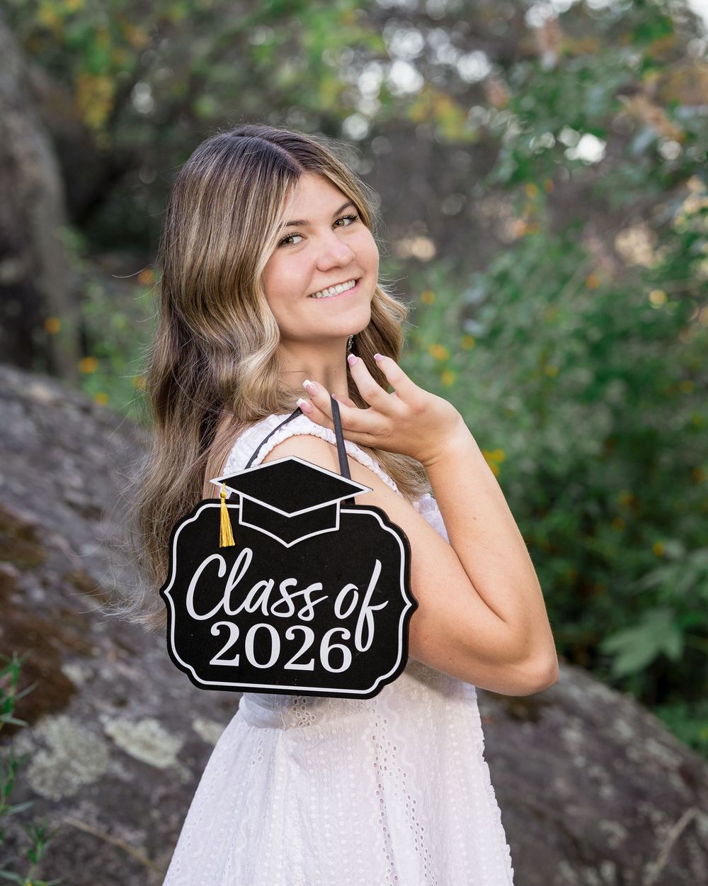 Smiling young woman holding a Class of 2026 graduation sign outdoors among green trees and rocks.