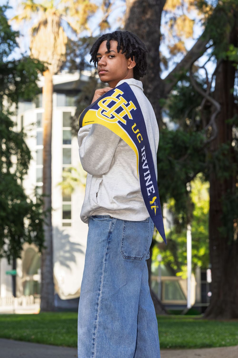 Student wearing UC Irvine sash draped over shoulder, posing outdoors on campus in jeans and hoodie.