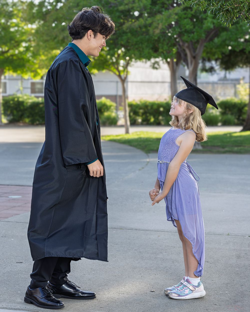 Graduate in black cap and gown shares a sweet moment with a young girl wearing a graduation cap outdoors.