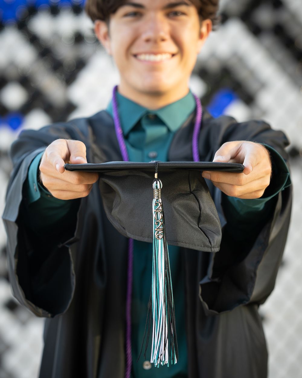 Smiling graduate in black gown holds mortarboard cap with green tassel toward camera at graduation ceremony.