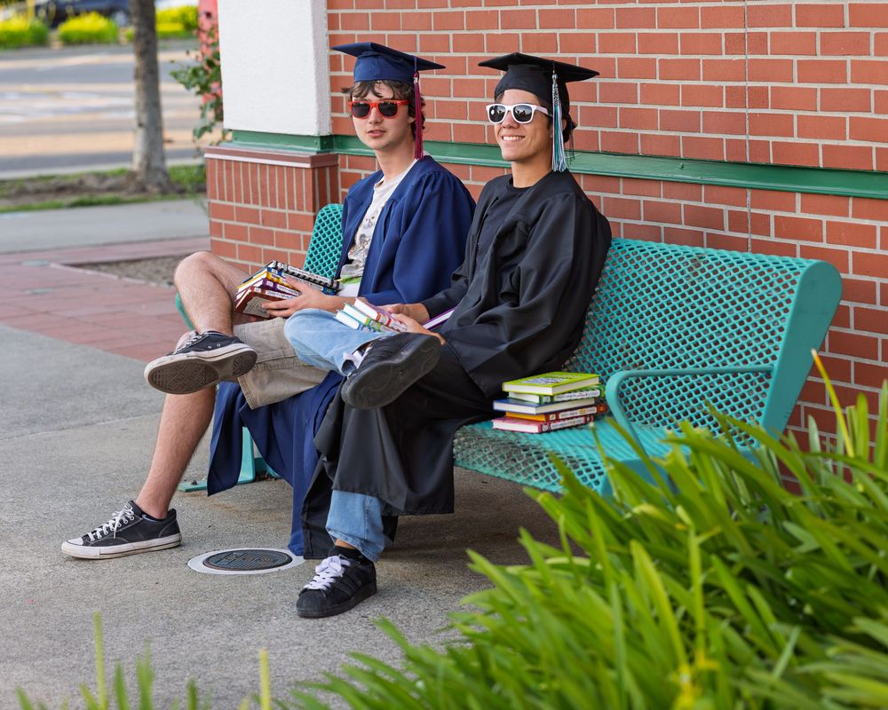 Two graduates in caps and gowns sit on a teal bench with stacked books, smiling outdoors on graduation day.
