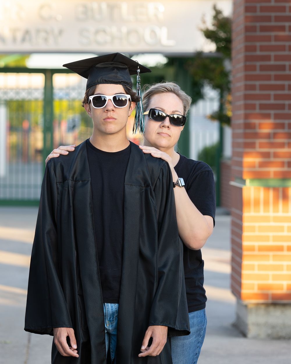 Graduate in black cap and gown poses with sunglasses beside proud parent outside school building.