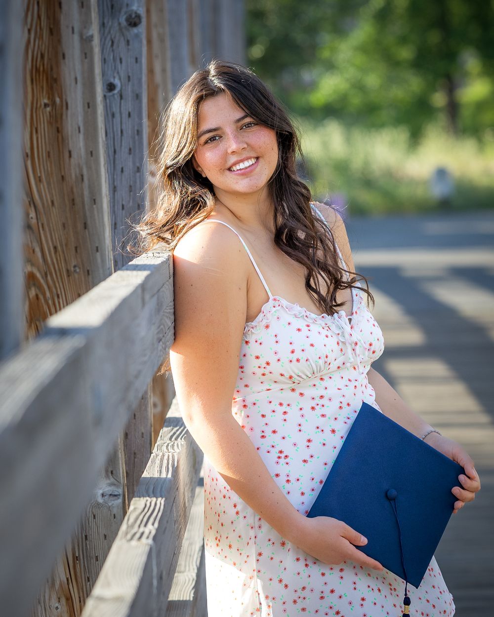 Smiling young woman in floral dress holding navy graduation cap and folder, leaning against wooden fence outdoors.