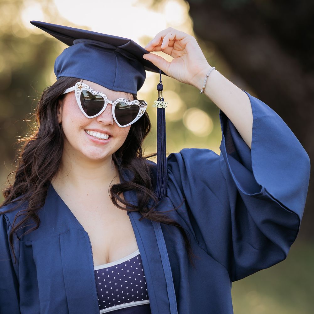 Smiling graduate in navy cap and gown wearing heart-shaped sunglasses, posing outdoors for graduation photos.